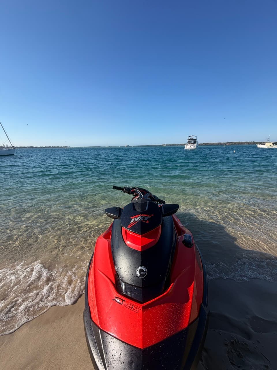 Red and black jet ski on a sandy beach overlooking clear blue water and boats.